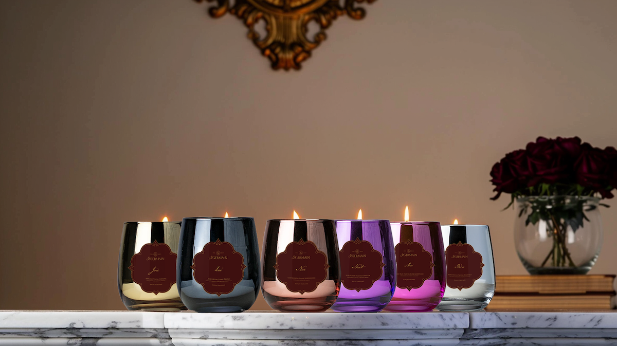 Five St.Germain candles in metallic vessels on a luxury marble table and flowers in the background.
